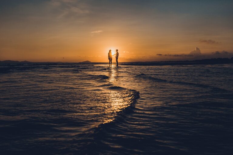 Instagram worthy photo of a couple standing in the distance at the beach with the sun setting between the couple