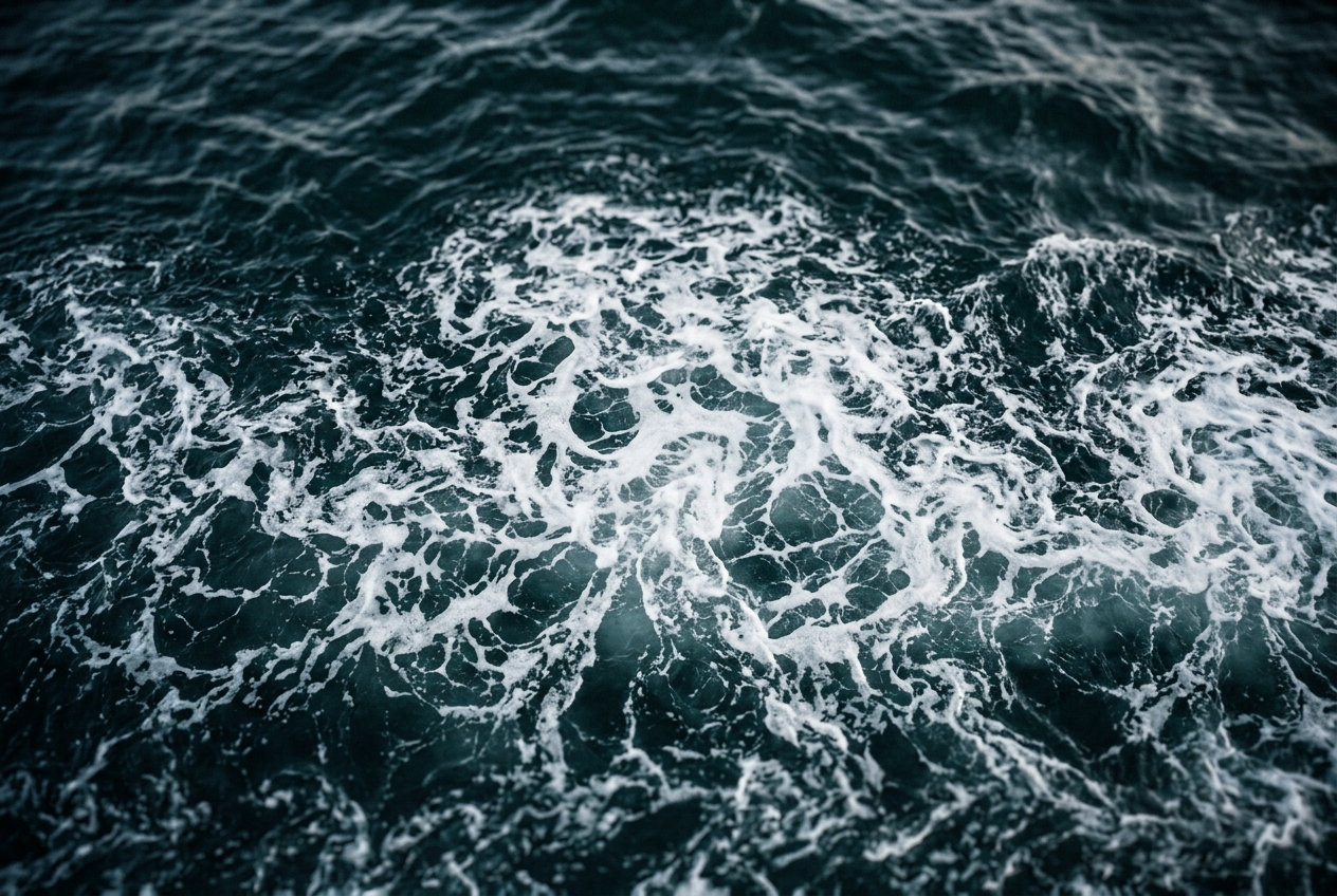 A band of white foam breaking into lace across dark blue-green water and dissolving at the edges